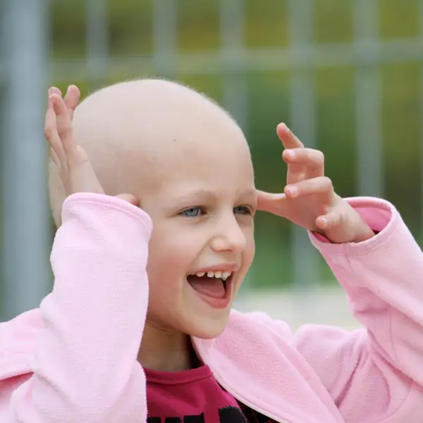 Smiling bald child wearing a pink jacket with hands raised near head outdoors.