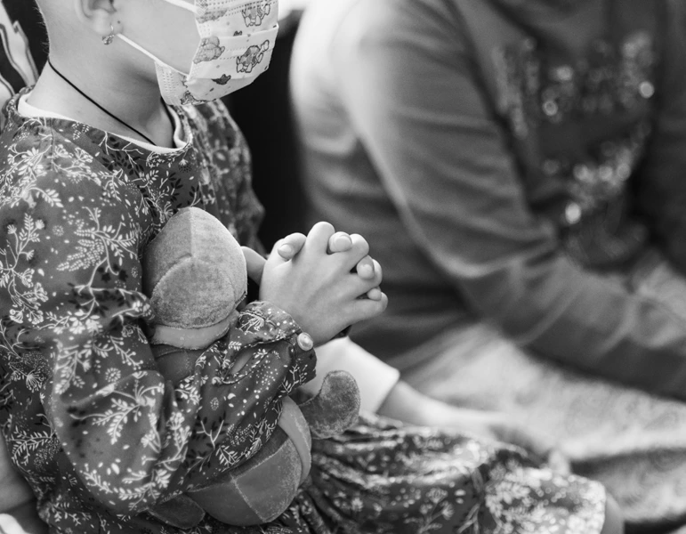 Child wearing a patterned mask and dress, clasping hands while holding a stuffed toy, seated next to another person.