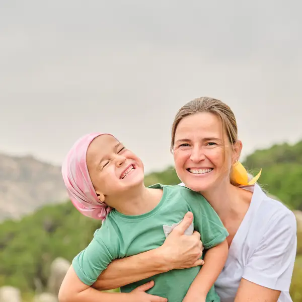 Smiling woman hugging a laughing child wearing a pink headscarf outdoors with blurred greenery in the background.