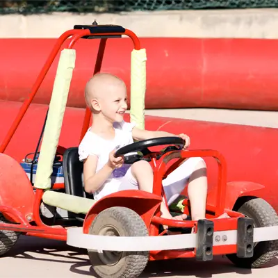 Young child with a bald head smiling while sitting and steering a red go-kart outdoors.