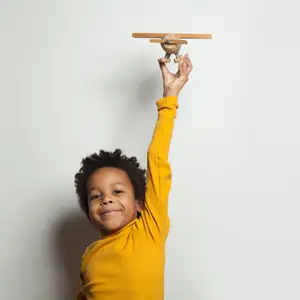 Smiling boy in a yellow shirt holding a wooden toy airplane above his head against a plain light background.