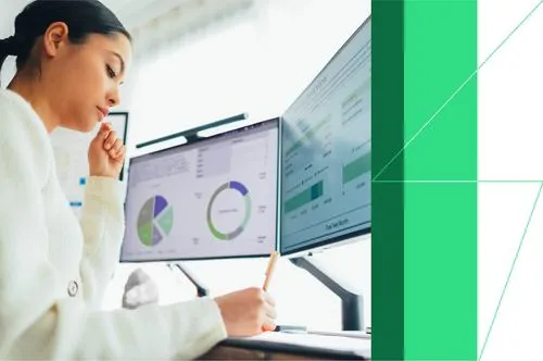 Woman analyzing data on two computer screens with pie charts and graphs for business finance insights.