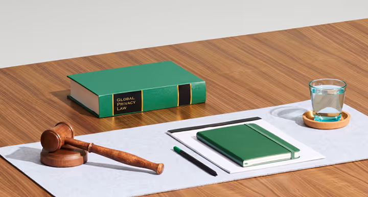 Green law book, gavel, notepad, pens, and glass on wooden desk with white paper background.