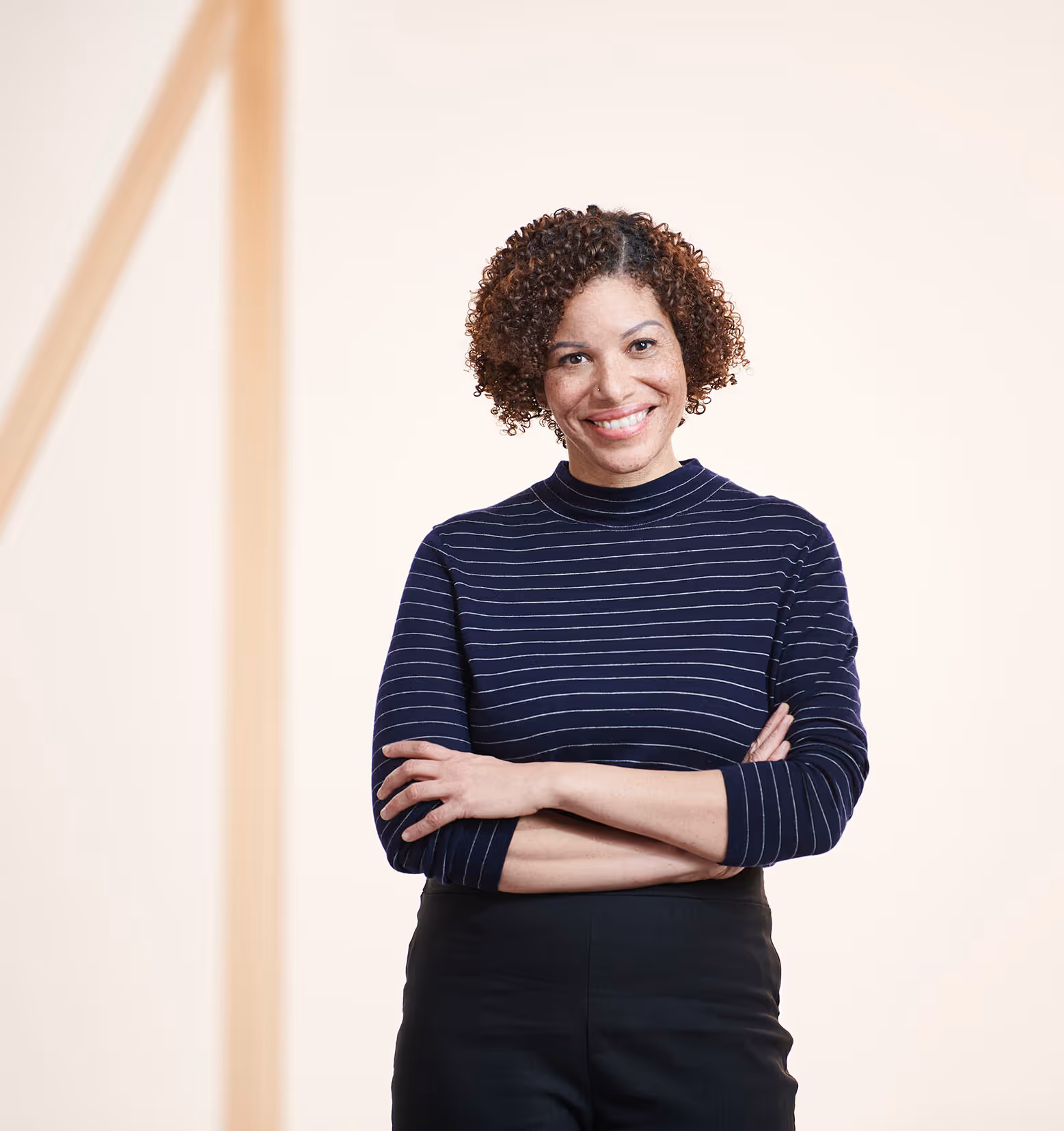 Woman with curly hair smiling and crossing arms, wearing striped shirt and black pants confidently.