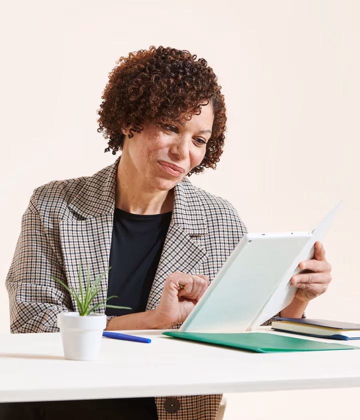 Businesswoman sitting at desk reviewing documents with a tablet and office supplies, curly hair.