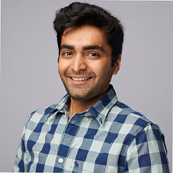 Smiling young man with dark hair wearing blue and green plaid button down shirt standing indoors.