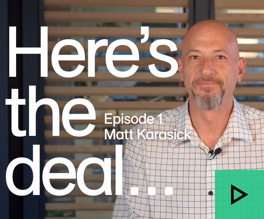 Bald man with checkered shirt and goatee standing in front of blinds and video play button.