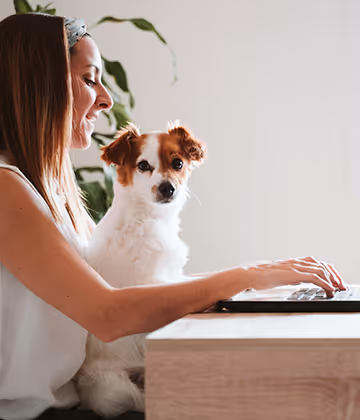 Woman working on laptop with Jack Russell Terrier dog sitting beside her on chair indoors.