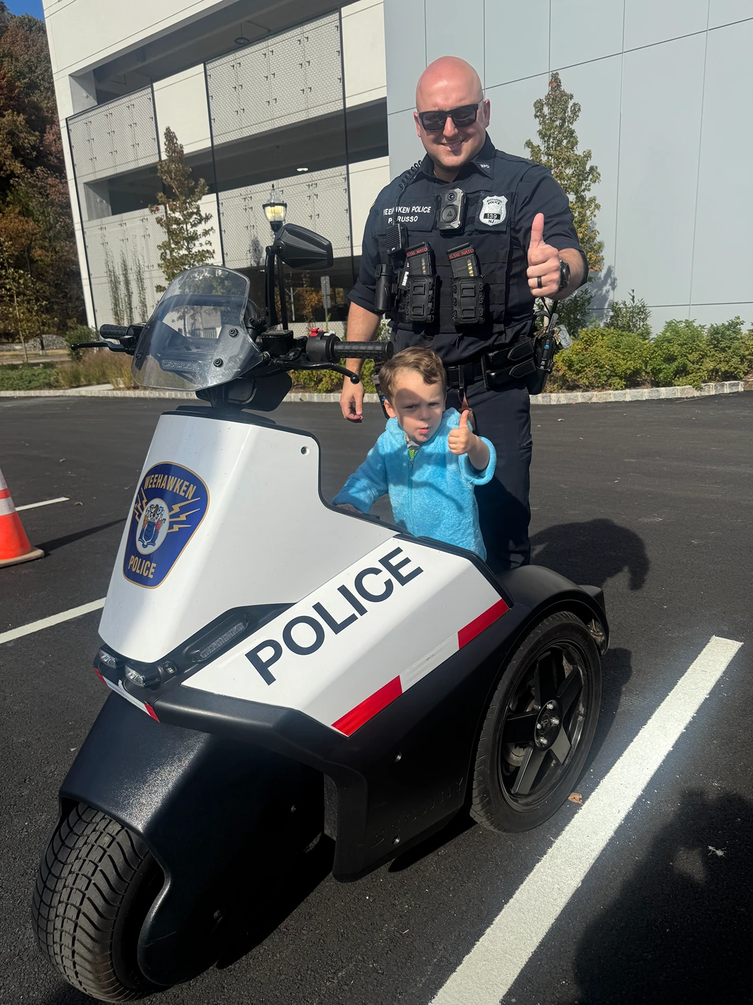 A smiling police officer stands next to a child giving a thumbs up while sitting on a police scooter in a parking lot. The police vehicle is marked with the word "POLICE" and has an emblem. Background features trees and a building.