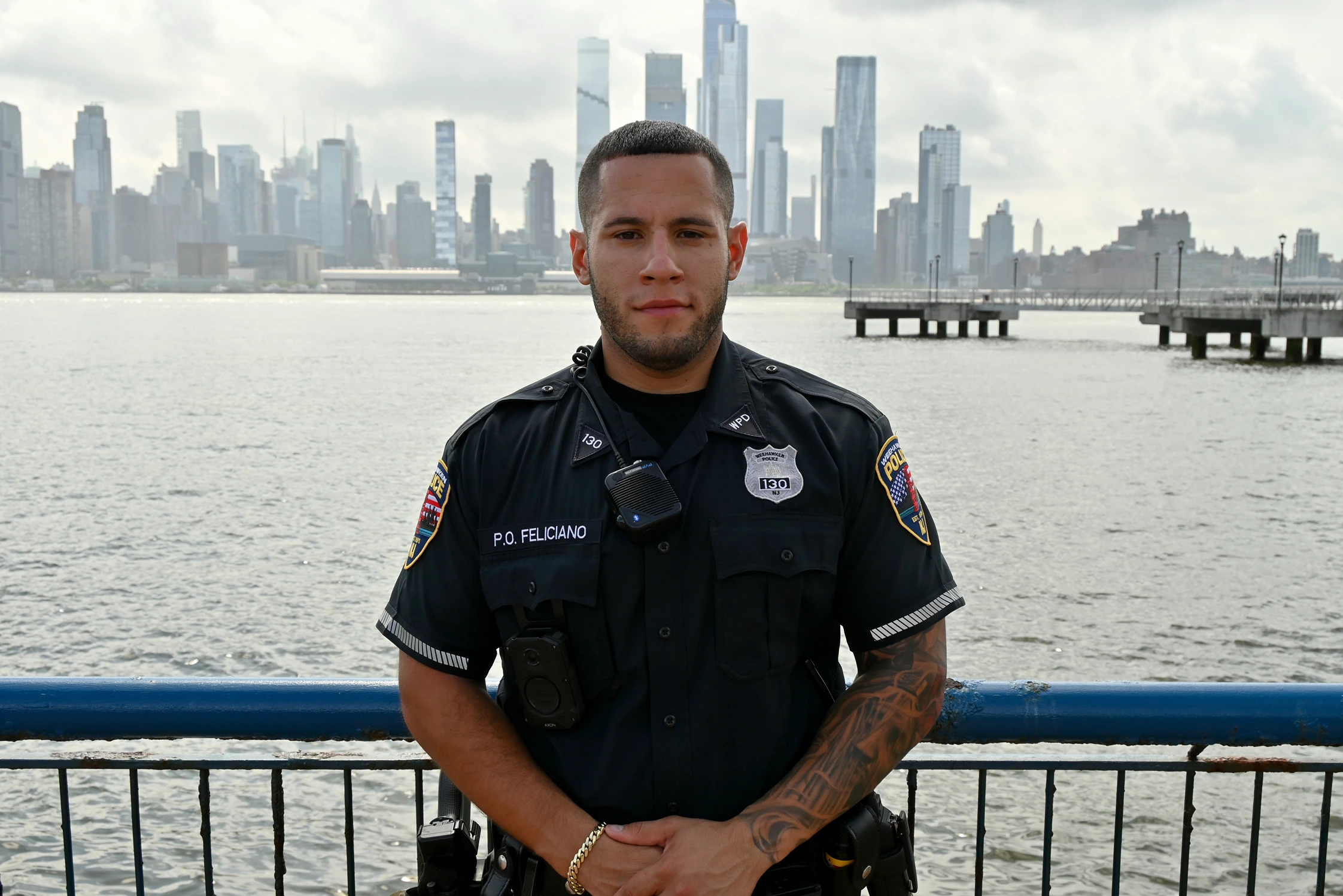 Police officer Will Feliciano stands by the waterfront, wearing a uniform with a badge, facing the skyline of a city in the background.