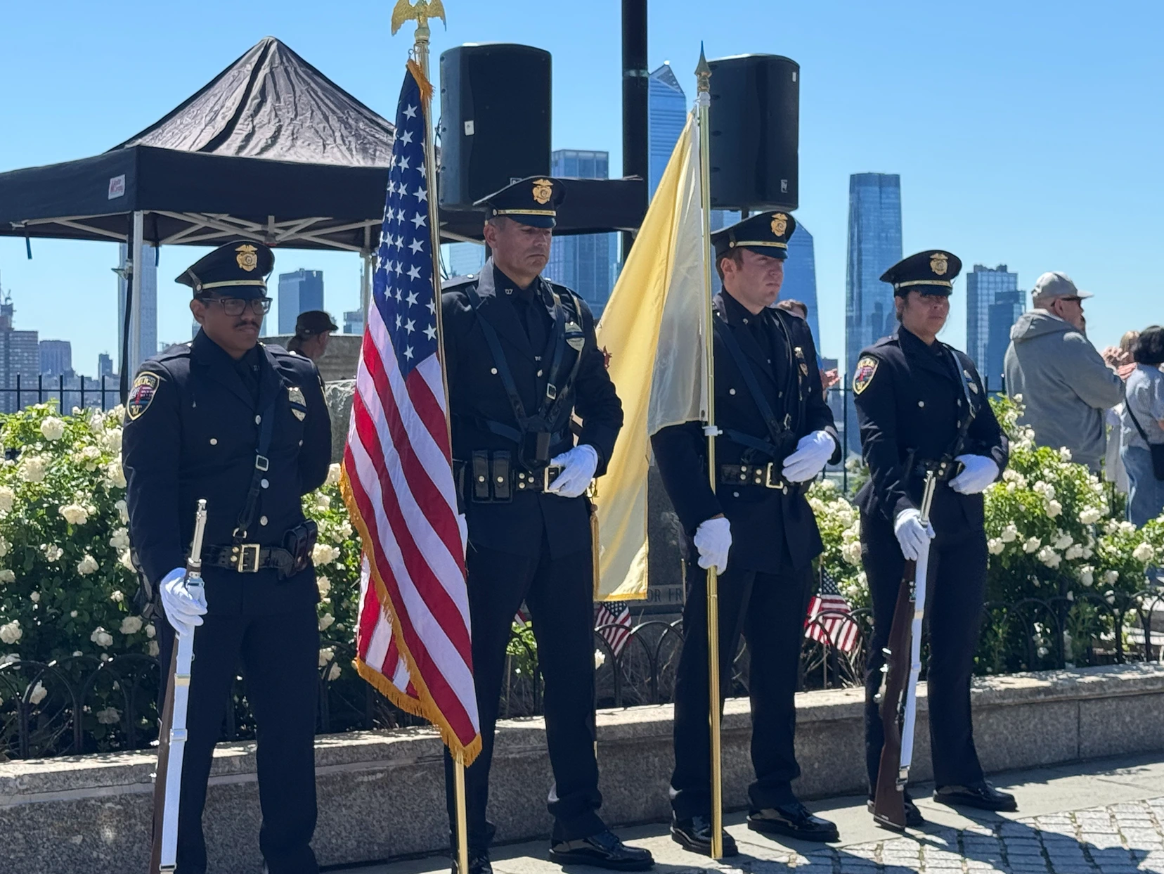 Four uniformed officers stand in formation, holding an American flag and a yellow flag, with city skyscrapers and a crowd in the background.
