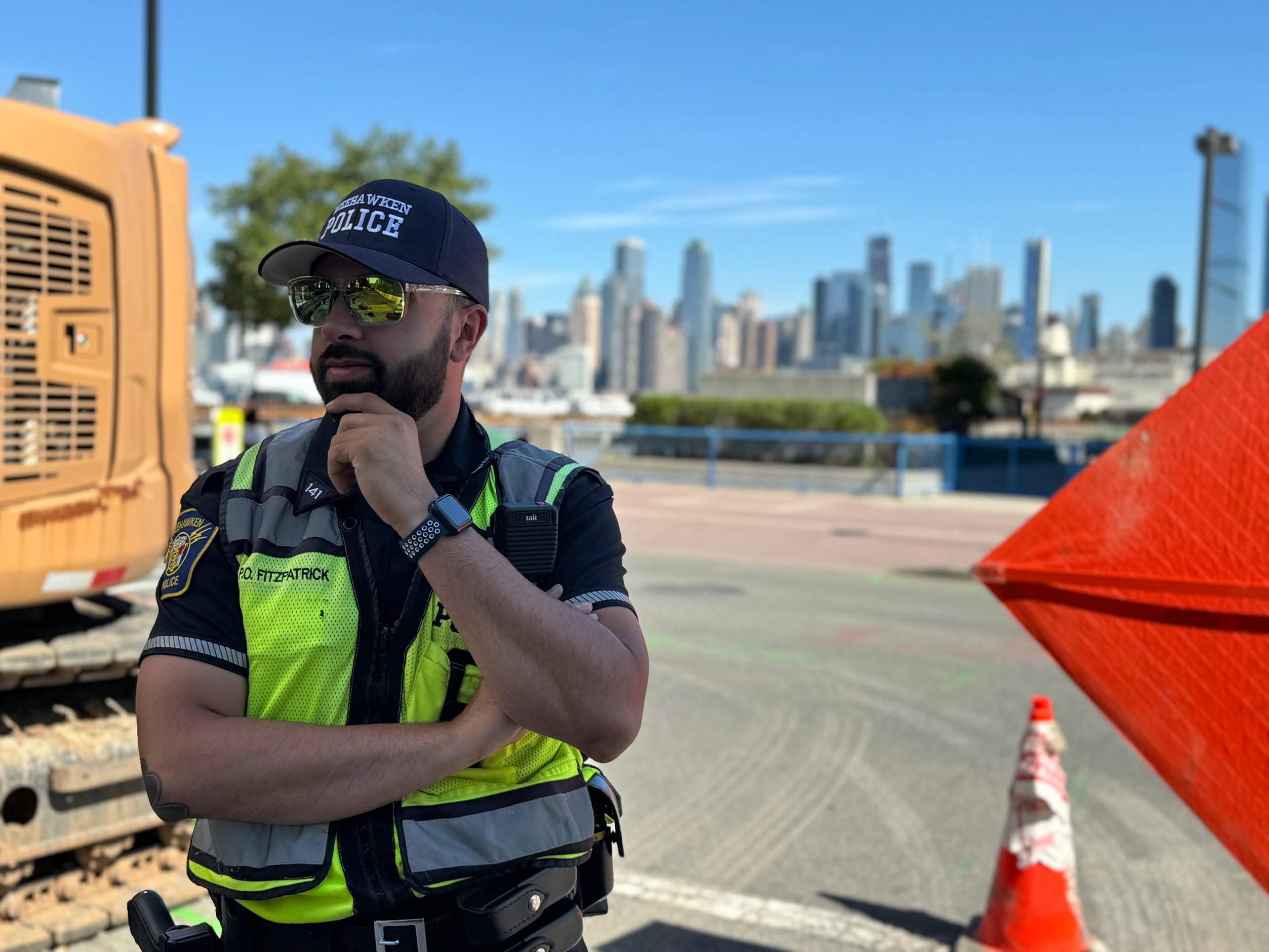 A group of police officers in uniforms standing on a street with trees and residential buildings in the background. One officer in the foreground wears sunglasses and a badge, while others engage in conversation. Several American flags are visible.