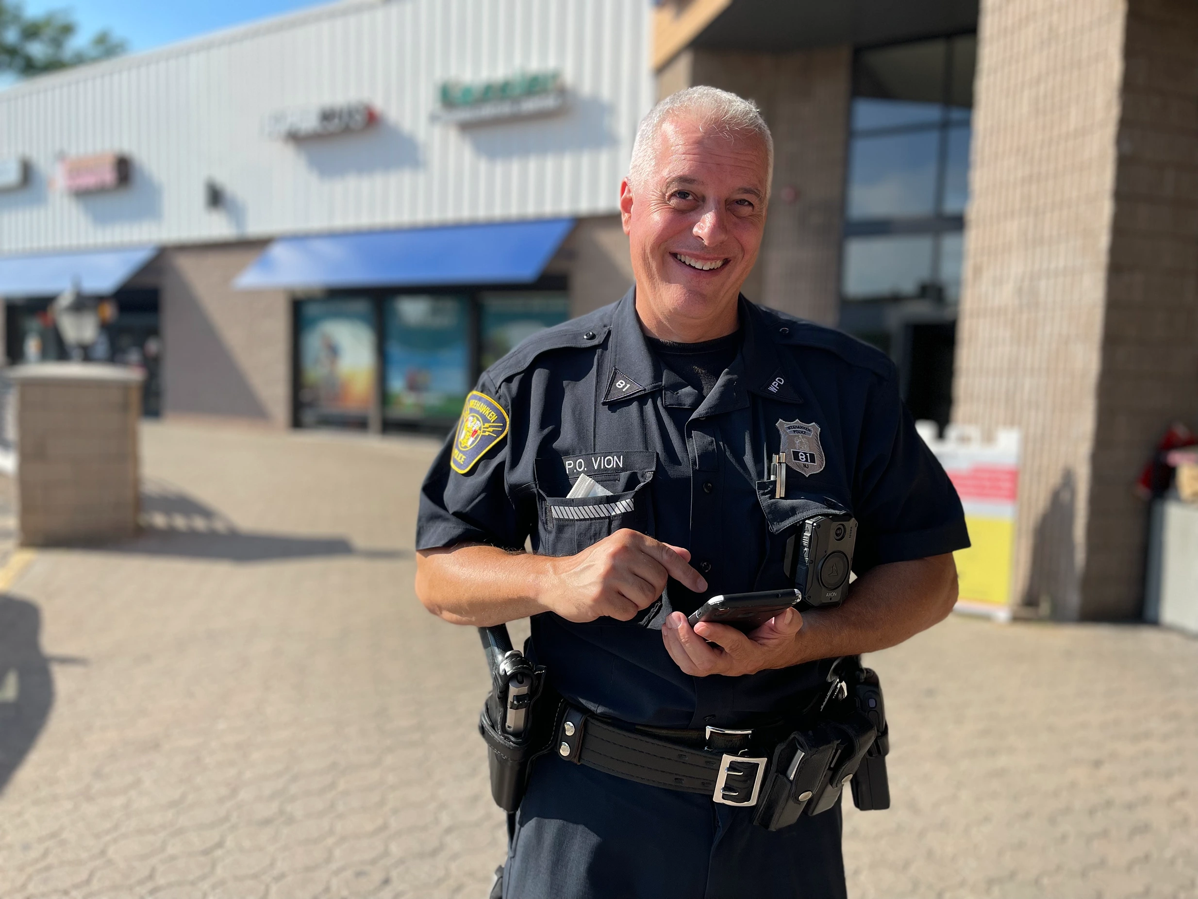 A group of police officers in uniforms standing on a street with trees and residential buildings in the background. One officer in the foreground wears sunglasses and a badge, while others engage in conversation. Several American flags are visible.