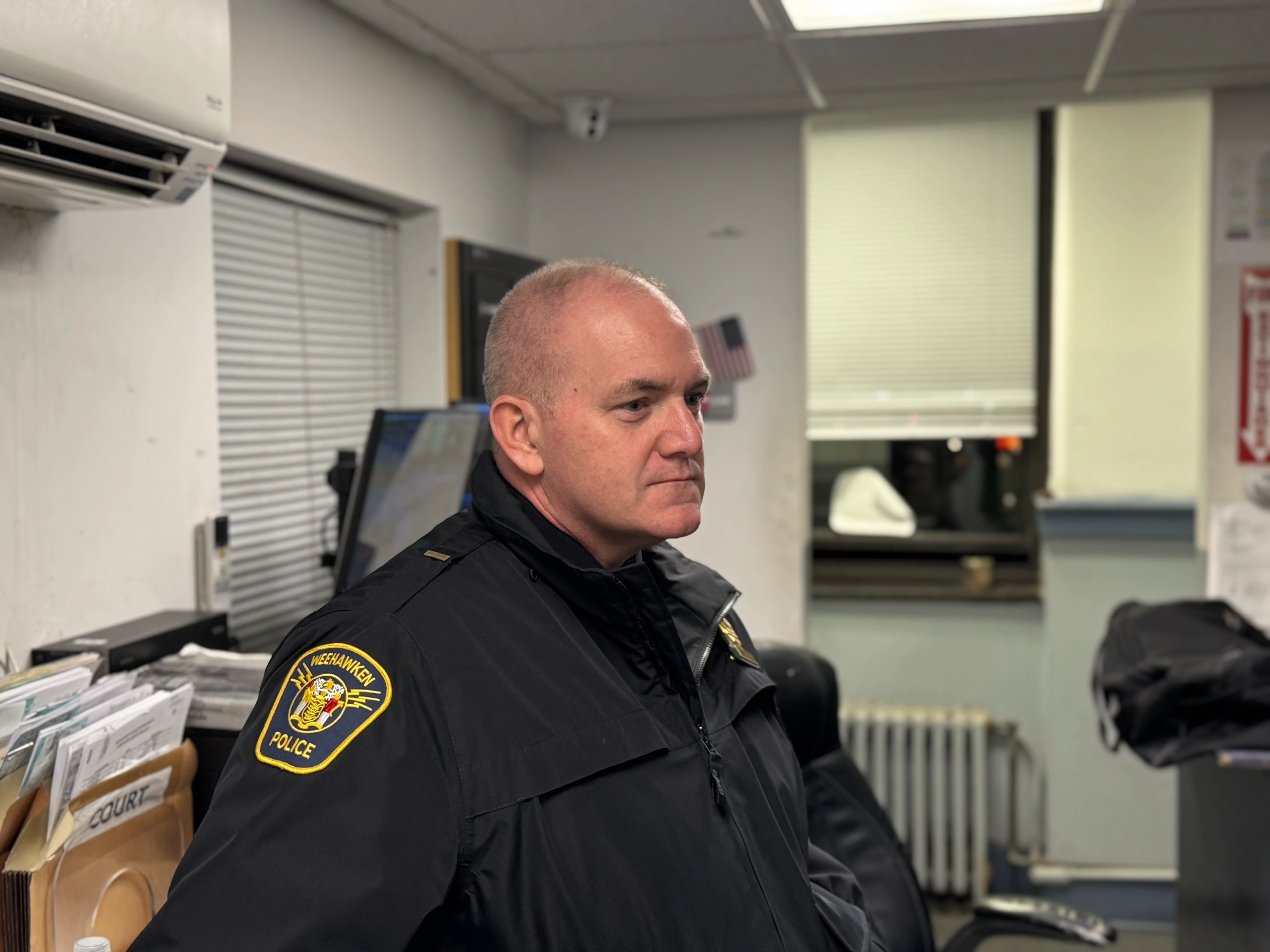 A group of police officers in uniforms standing on a street with trees and residential buildings in the background. One officer in the foreground wears sunglasses and a badge, while others engage in conversation. Several American flags are visible.