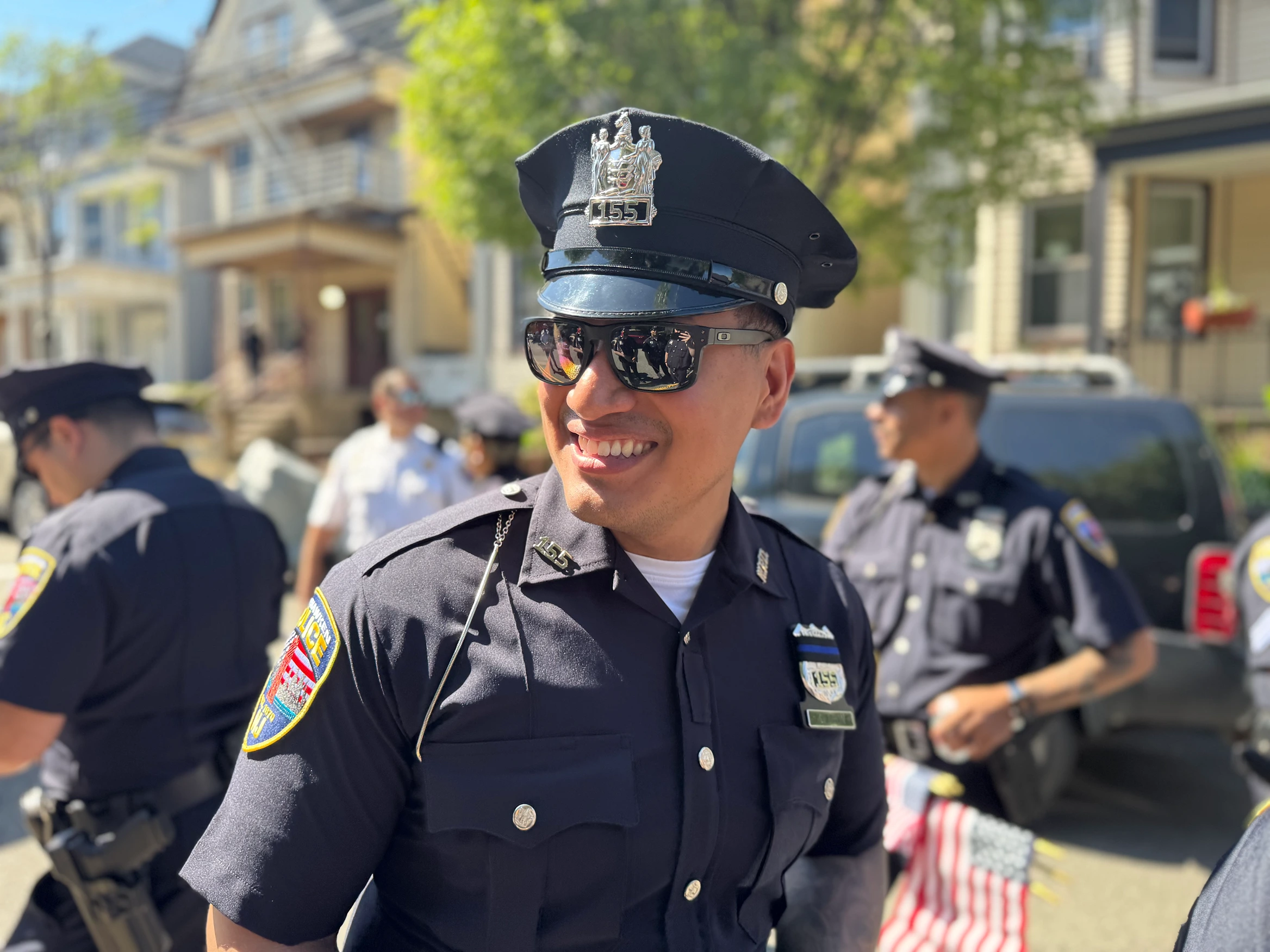 A group of police officers in uniforms standing on a street with trees and residential buildings in the background. One officer in the foreground wears sunglasses and a badge, while others engage in conversation. Several American flags are visible.
