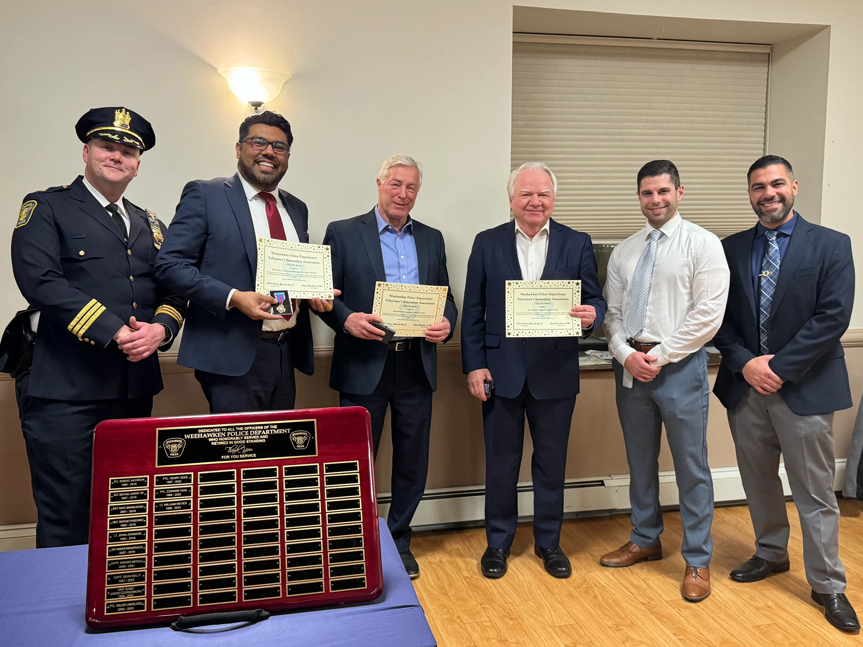 A group of police officers in uniforms standing on a street with trees and residential buildings in the background. One officer in the foreground wears sunglasses and a badge, while others engage in conversation. Several American flags are visible.