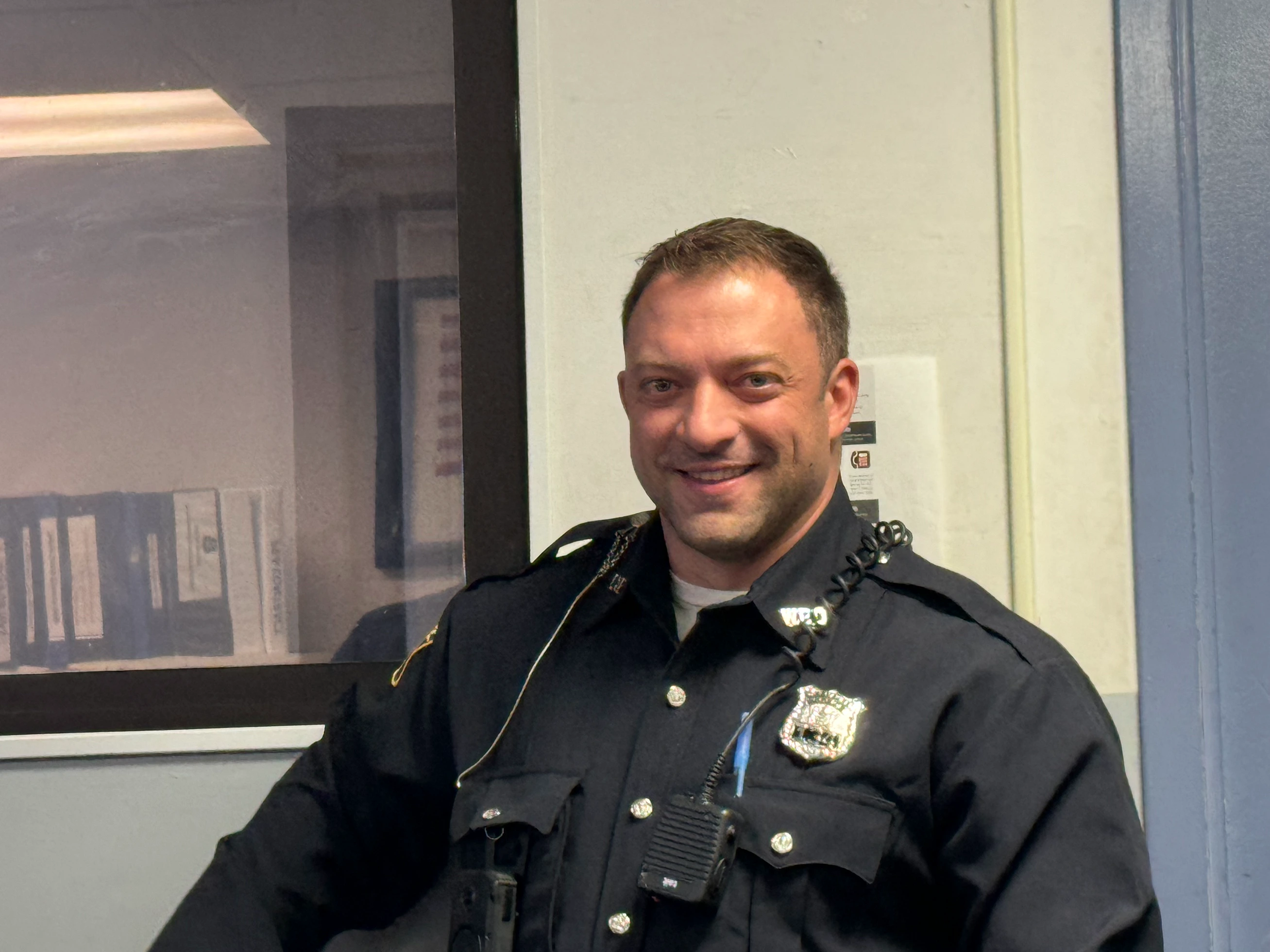 A group of police officers in uniforms standing on a street with trees and residential buildings in the background. One officer in the foreground wears sunglasses and a badge, while others engage in conversation. Several American flags are visible.