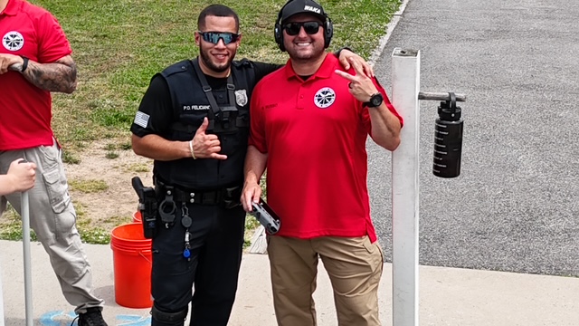 A police officer holding a chicken outside a house with flowers in the foreground. There is a police vehicle parked nearby, and another officer can be seen in a window.