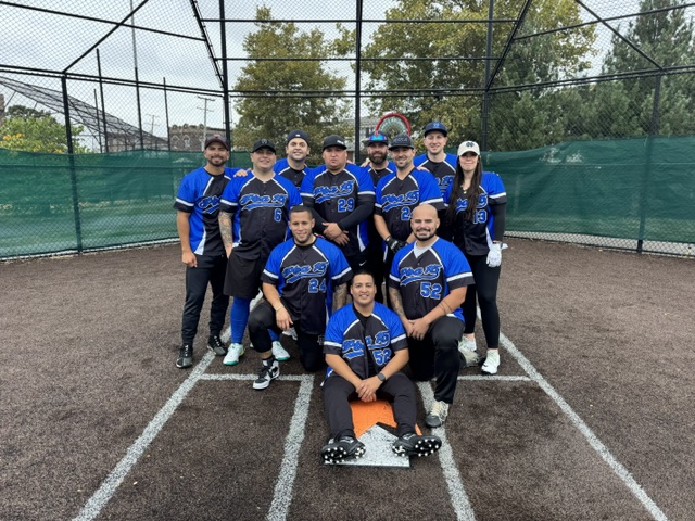 A group of police officers in uniforms standing on a street with trees and residential buildings in the background. One officer in the foreground wears sunglasses and a badge, while others engage in conversation. Several American flags are visible.