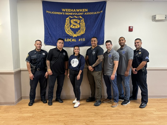 A group of police officers in bright yellow jackets and hats posing for a selfie at night, with a fenced area and a vehicle in the background.