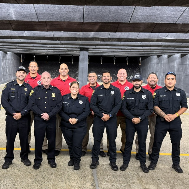 A group of police officers in uniforms standing on a street with trees and residential buildings in the background. One officer in the foreground wears sunglasses and a badge, while others engage in conversation. Several American flags are visible.