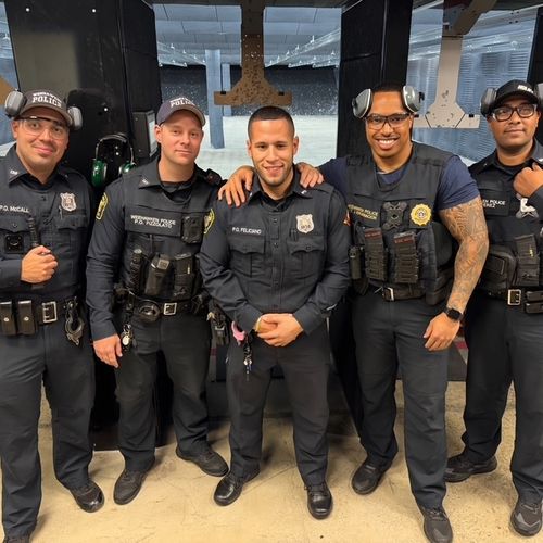 Two police officers in uniform, smiling and posing for a photo, with a city skyline in the background. The scene includes trees and a crowd of people, indicating a community event.
