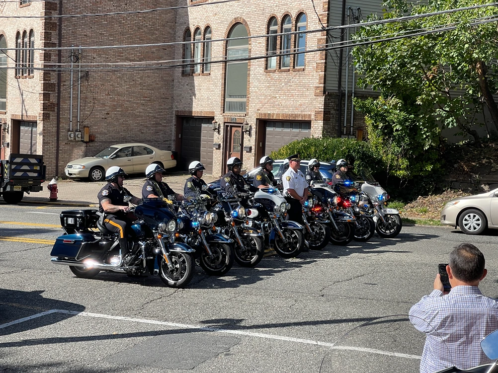 A line of police officers on motorcycles stands in front of a brick building, with one officer in a white uniform and a spectator taking a photo.