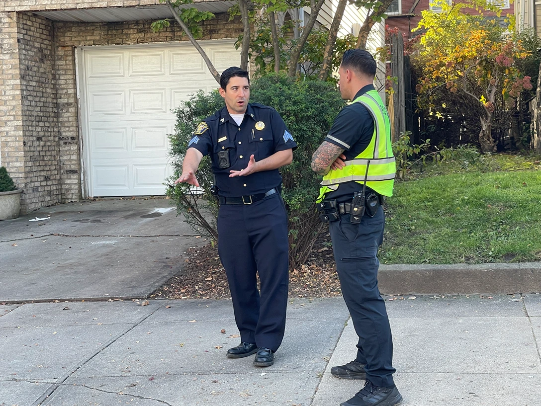 Two uniformed officers engage in conversation in front of a residential garage, surrounded by greenery and autumn foliage.