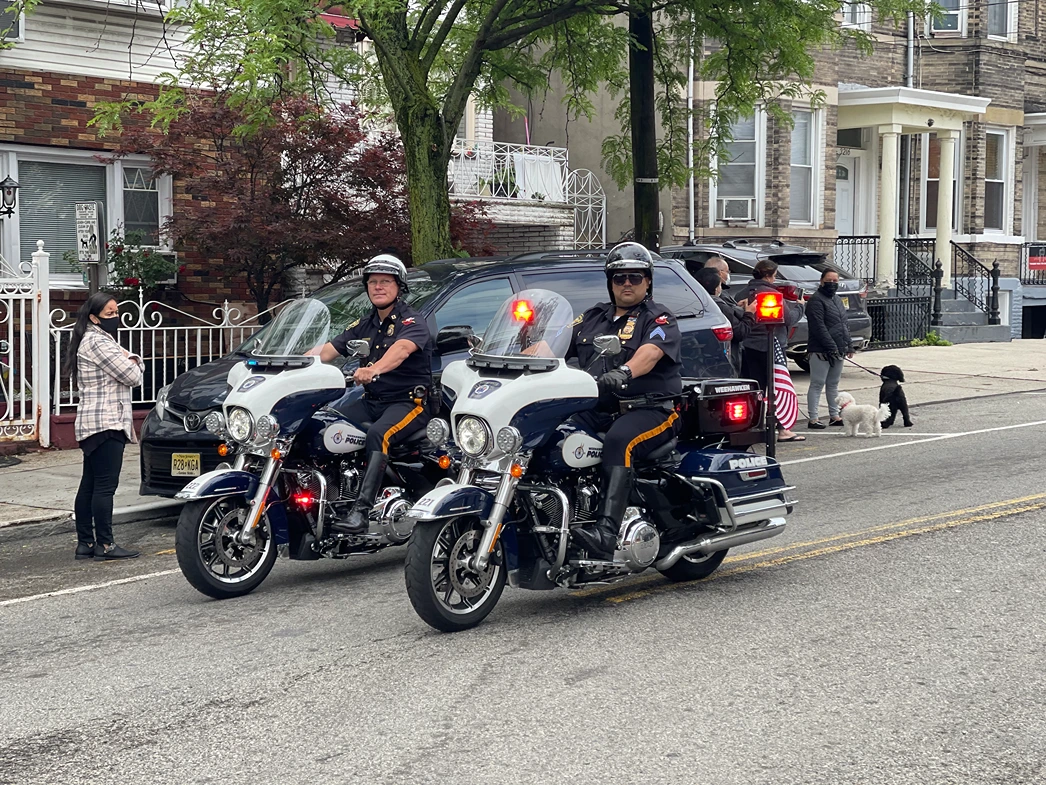 Two police officers on motorcycles talk to a woman on the sidewalk, while a couple walks dogs nearby in a residential area.