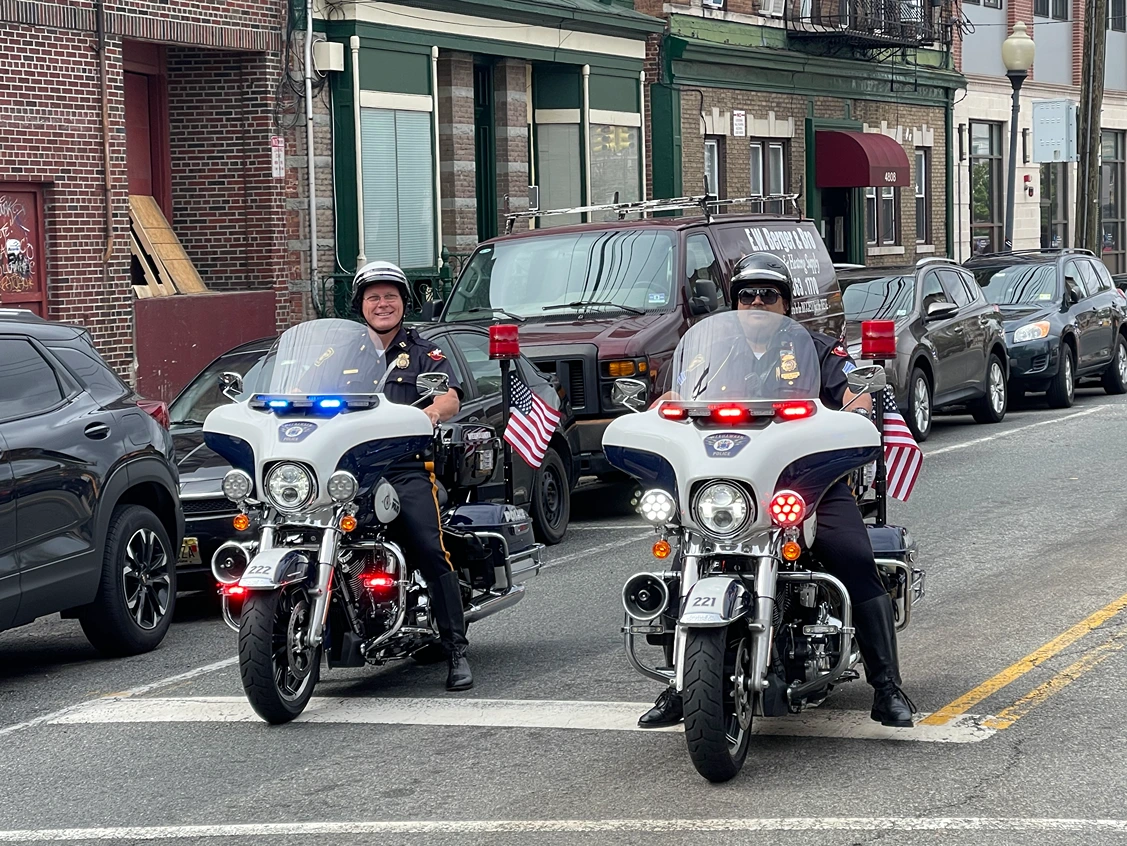 Two police officers on motorcycle units are parked at an intersection, displaying lights and American flags, with parked cars in the background.