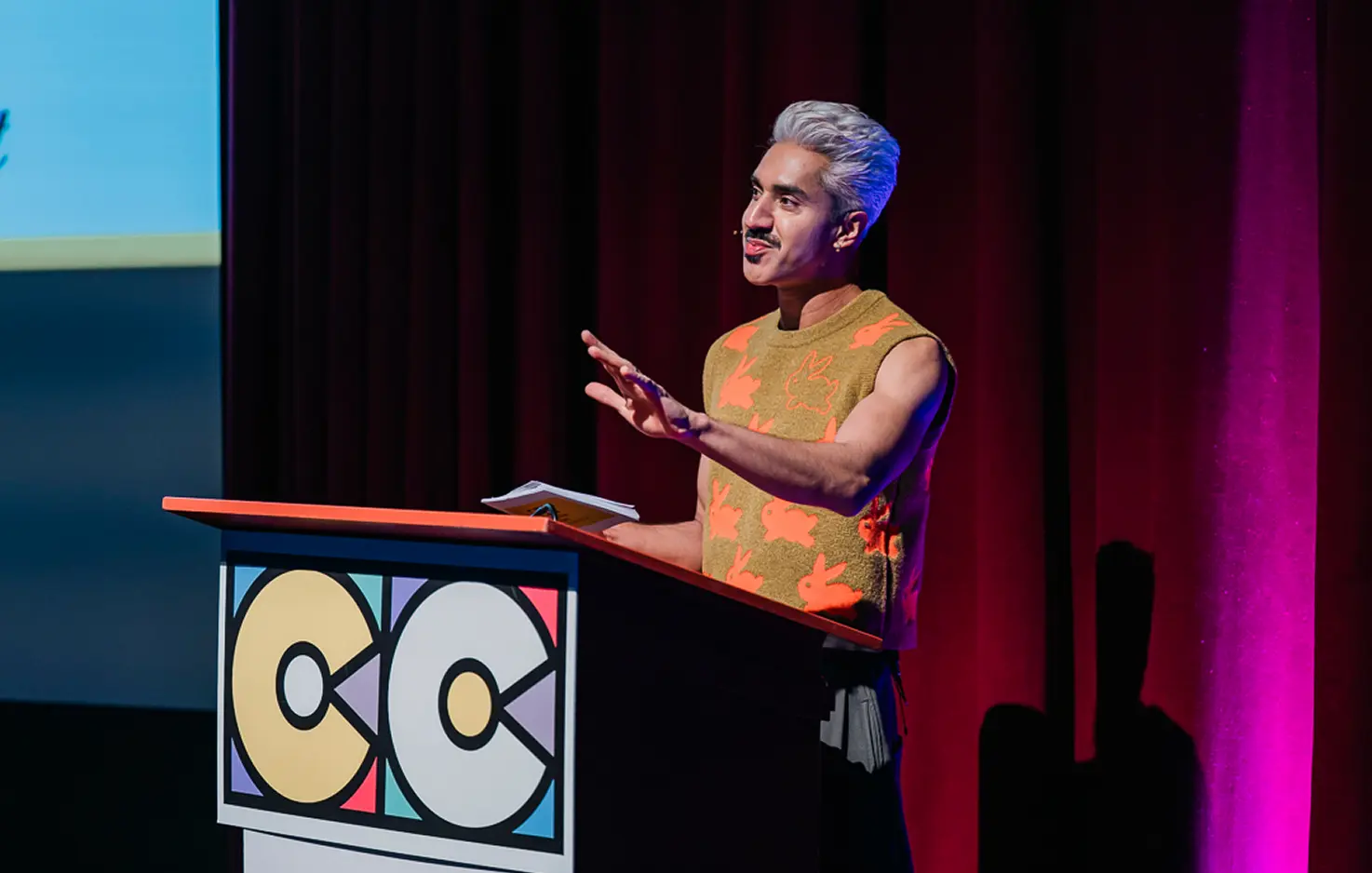 Person with silver hair and a mustard sweater with orange bunny patterns speaking at a podium with a CC logo, against a dark red curtain.