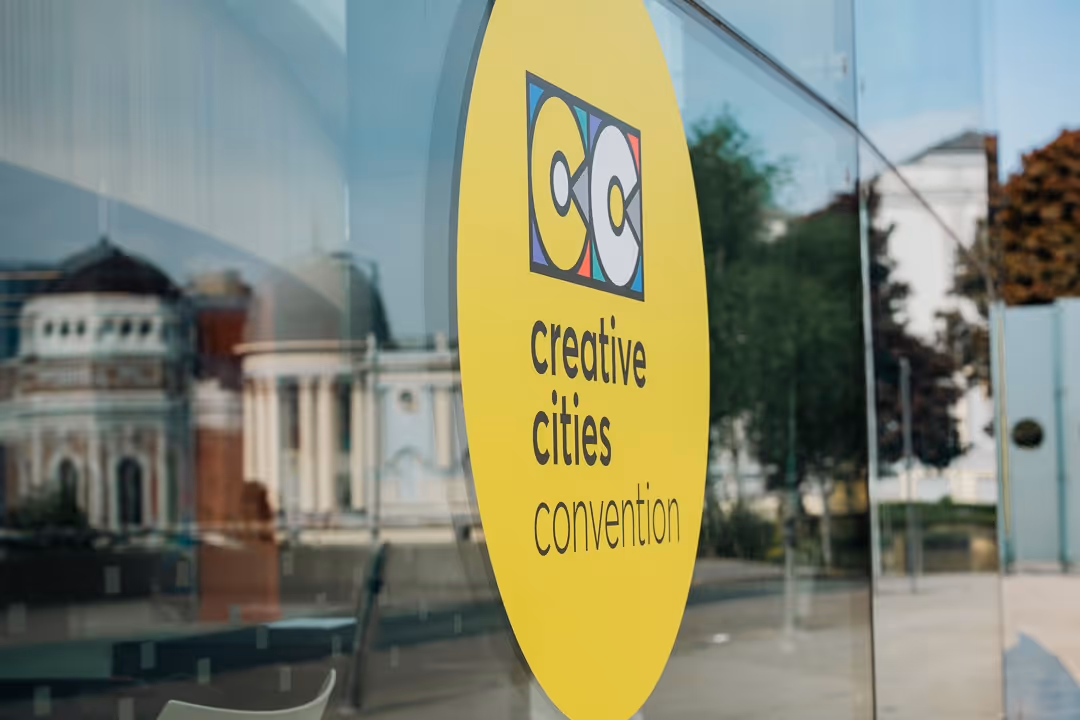 Yellow circular sign on glass window displaying the logo and text 'Creative Cities Convention' with blurred urban buildings reflected in the glass.