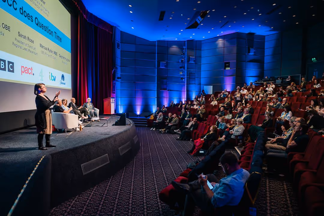 Panel discussion with four speakers seated on stage and a sign language interpreter standing in front of an audience in a theater with blue lighting.
