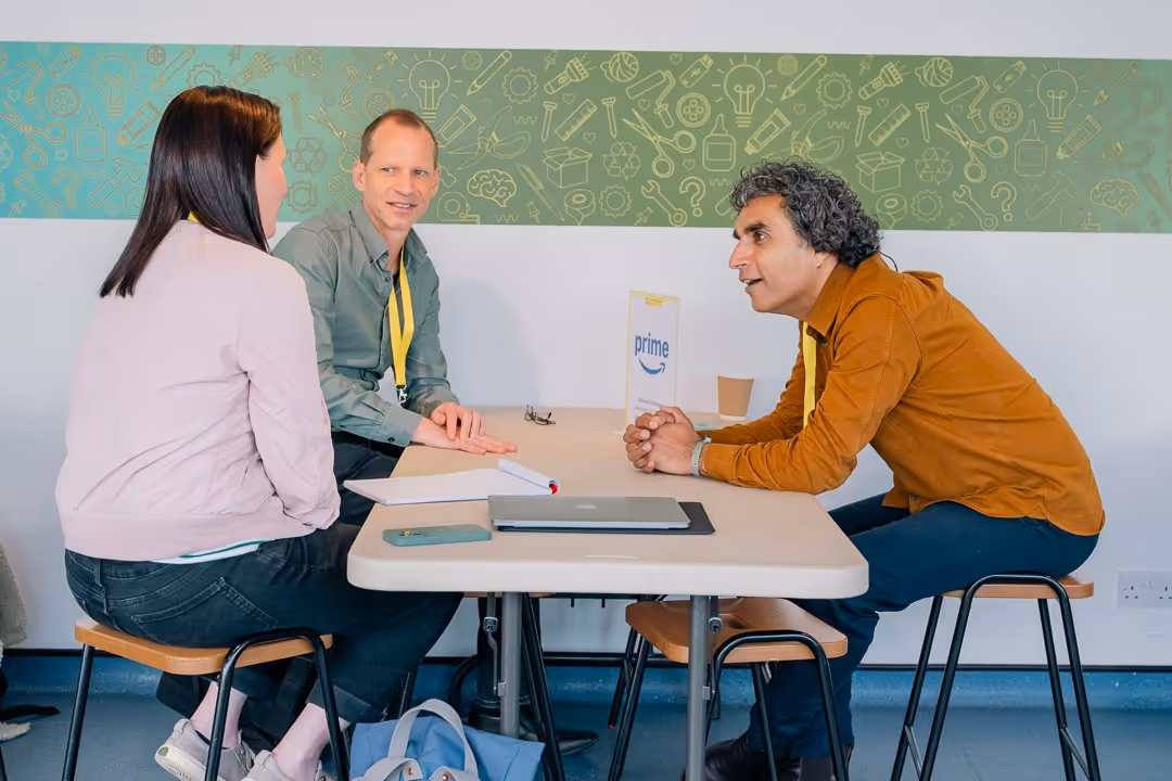 Three people having a discussion while sitting on stools around a table with a laptop and phone on it, against a wall with green patterned decor.