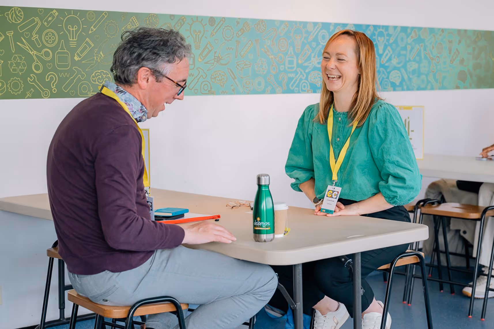 Two people wearing conference badges laughing and talking across a table in a meeting room.