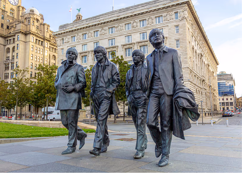 Statue of The Beatles walking in a row on a city sidewalk with historic buildings and trees in the background.