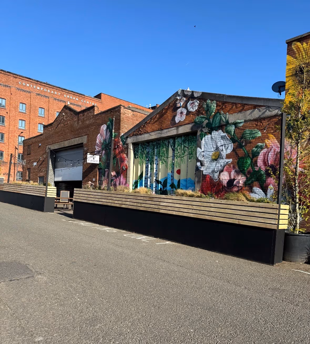 Brick building with colorful floral mural and a sign reading 'The Furnace' under clear blue sky.