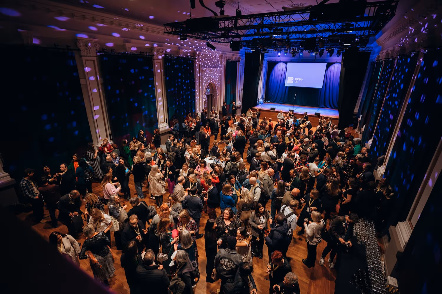 Large crowd mingling in a decorated event hall with purple lighting and a stage at the front displaying a screen.