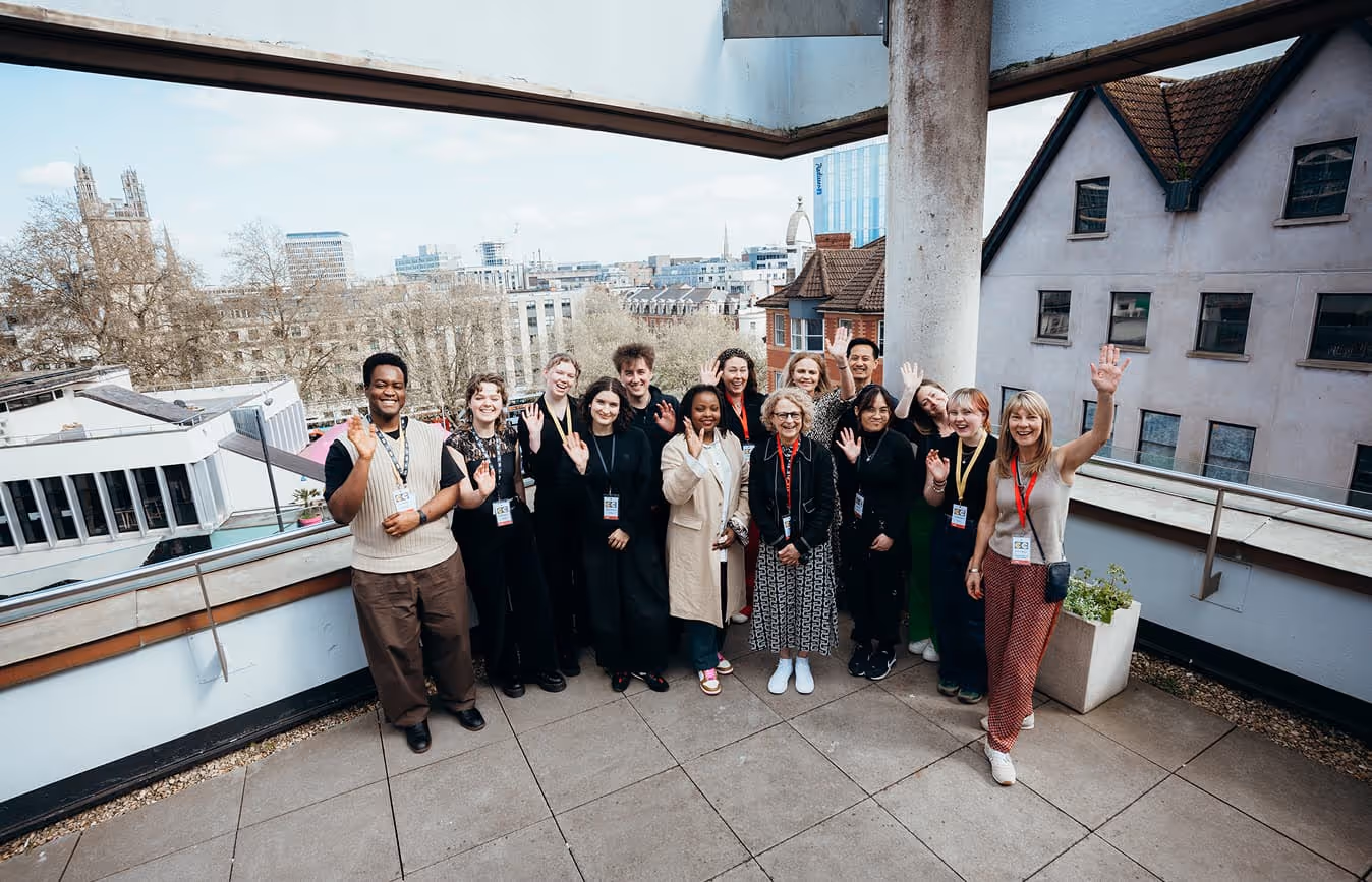 Group of diverse people standing on a rooftop terrace waving and smiling at the camera with urban buildings in the background.
