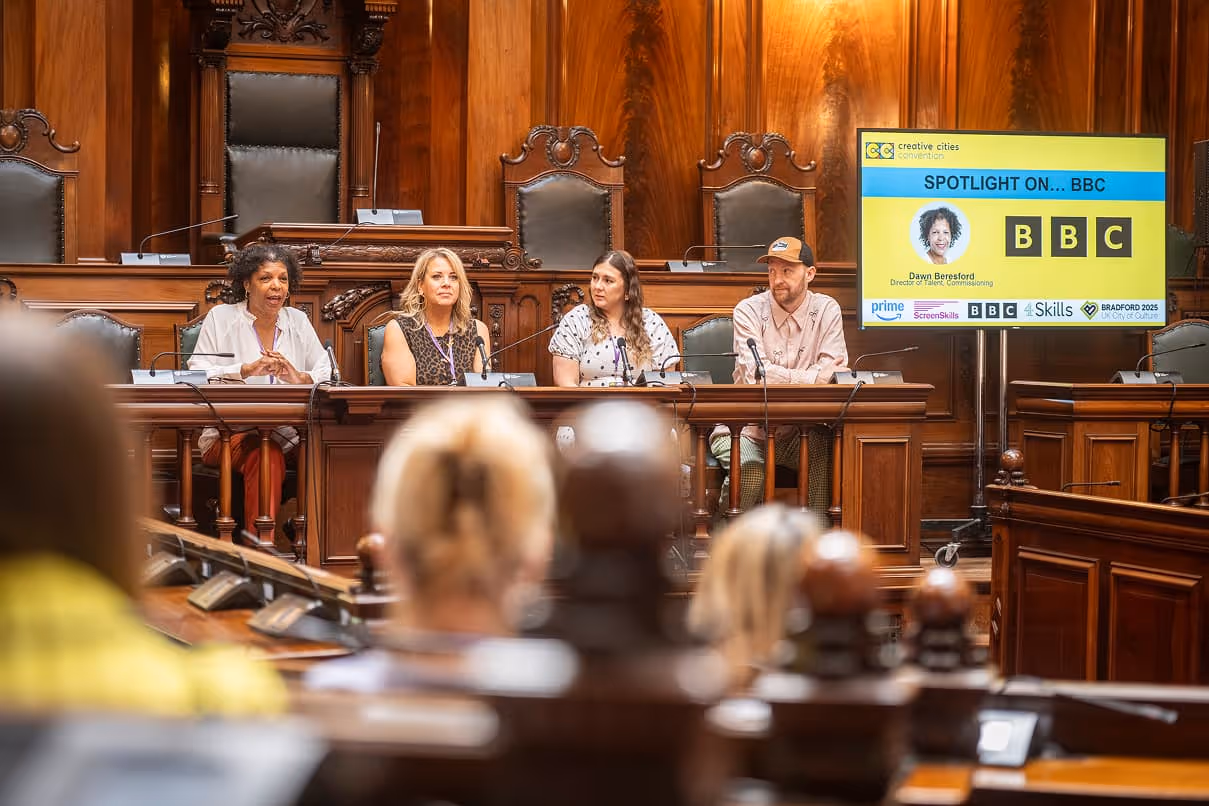 Panel of four people speaking at a conference table in a wood-paneled room with a screen displaying 'Spotlight on... BBC' in the background.