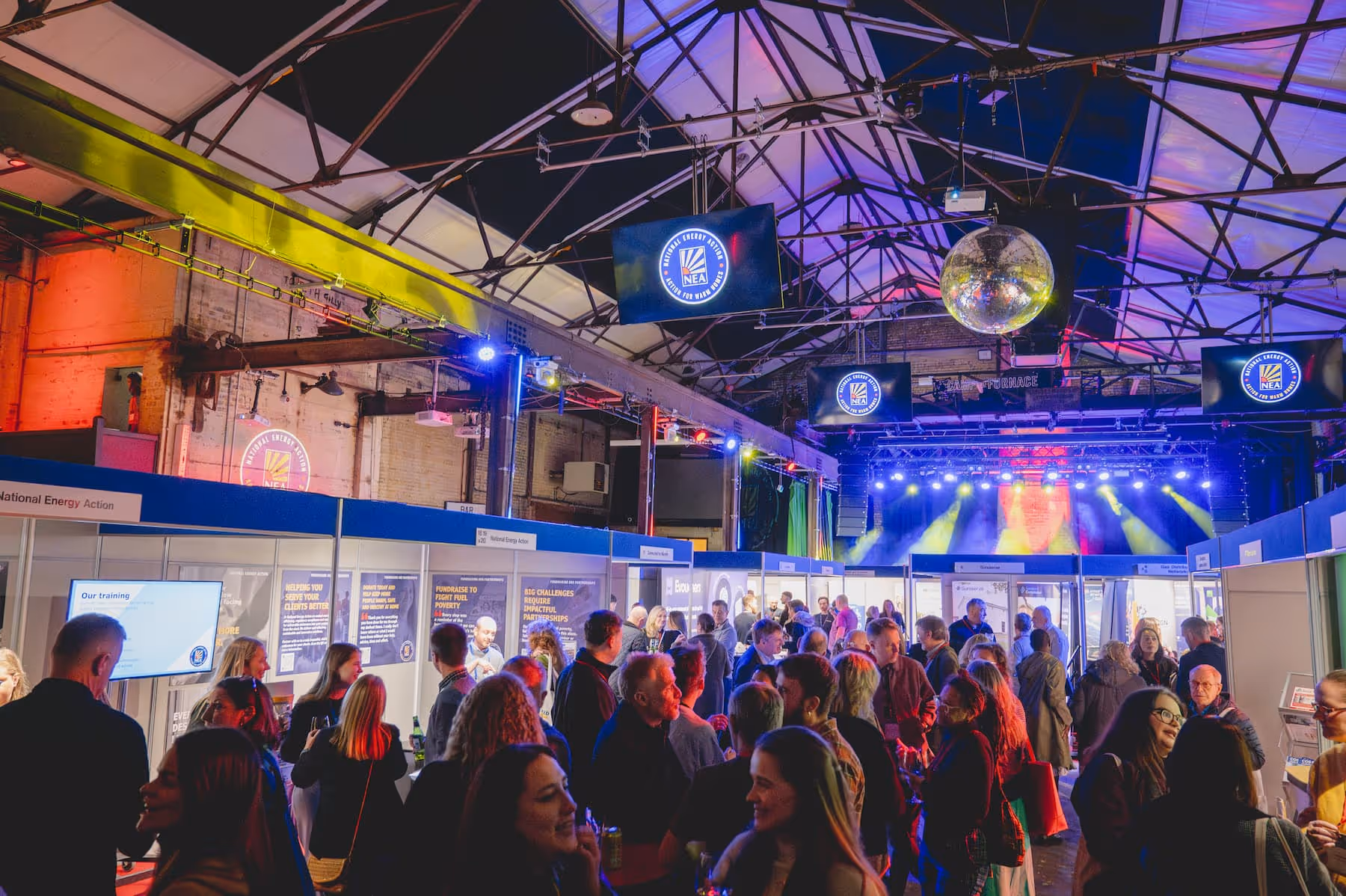 Crowded indoor event with colorful lighting, booths, and multiple screens displaying the National Energy Action logo.