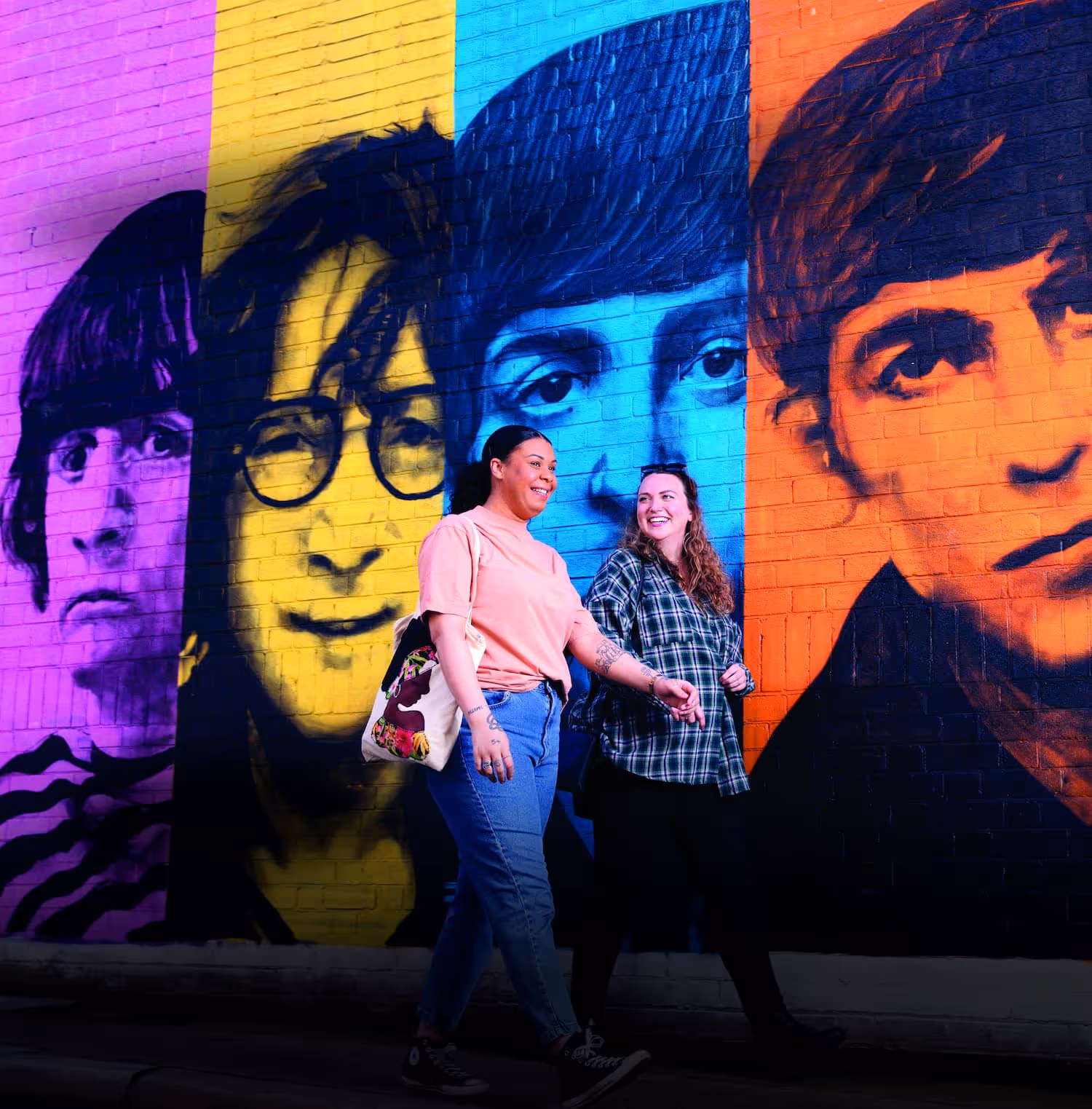 Two women smiling and walking past a colorful mural of four faces painted on a brick wall in purple, yellow, blue, and orange panels.