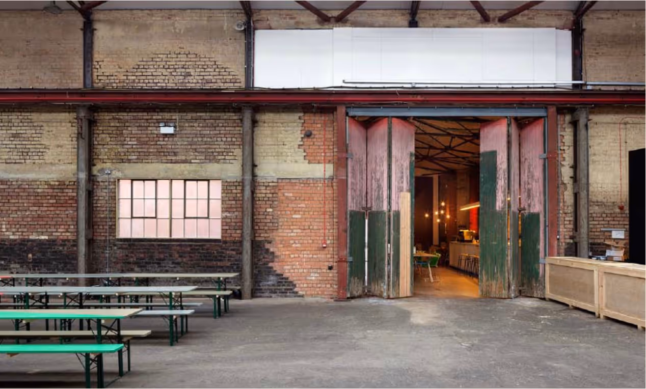 Interior of a rustic industrial-style space with large folding wooden doors partially open, green and beige benches, exposed brick walls, and warm lighting inside.