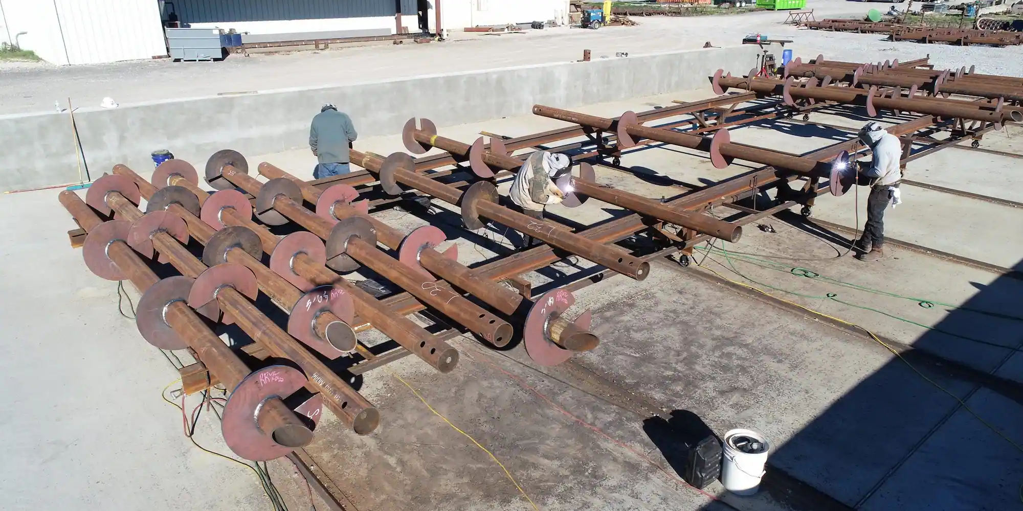 Two welders working on large metal pipes with flange plates arranged on the ground in an outdoor industrial area.