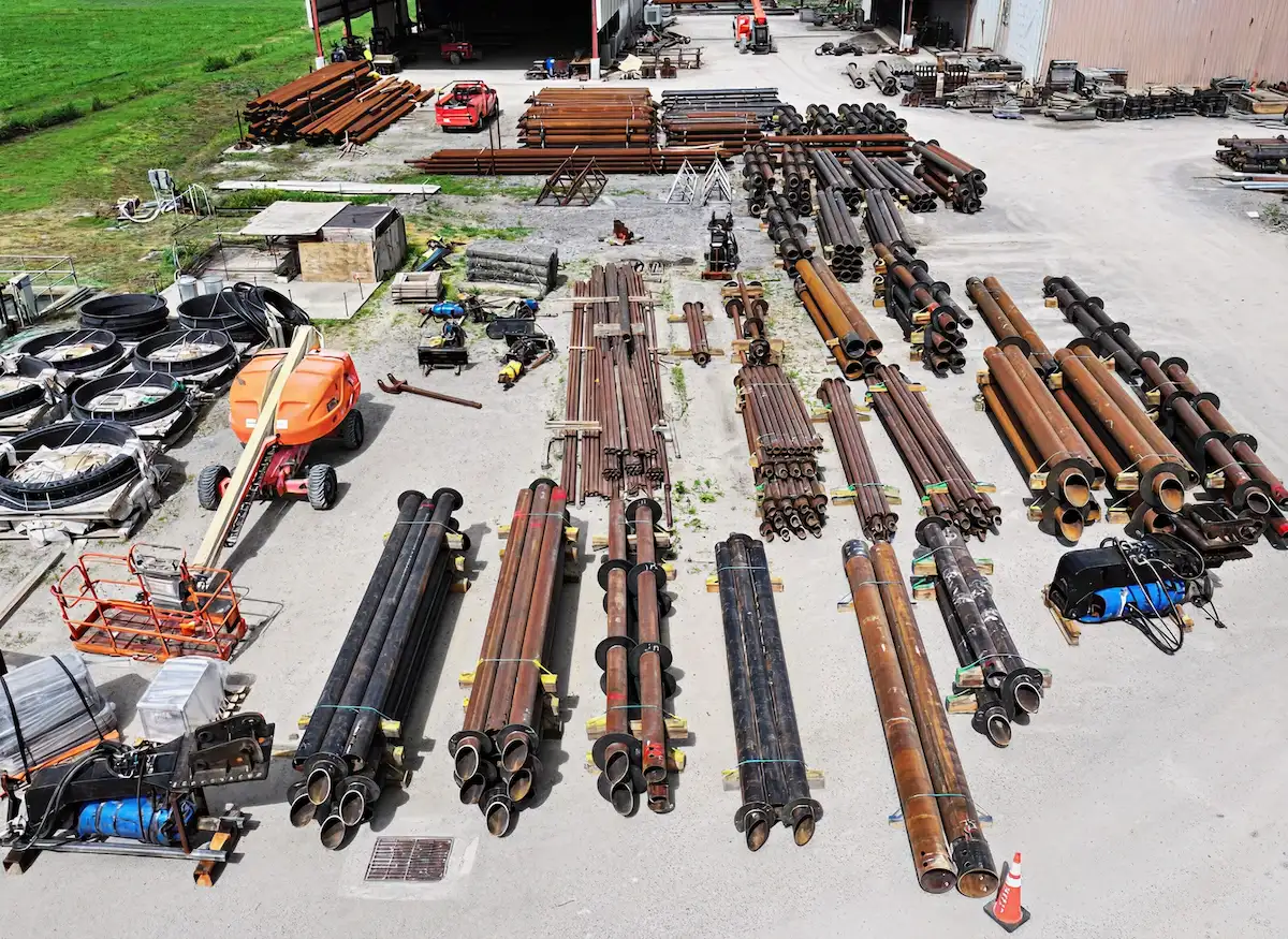 Industrial yard with organized stacks of large metal pipes and construction equipment on concrete ground.