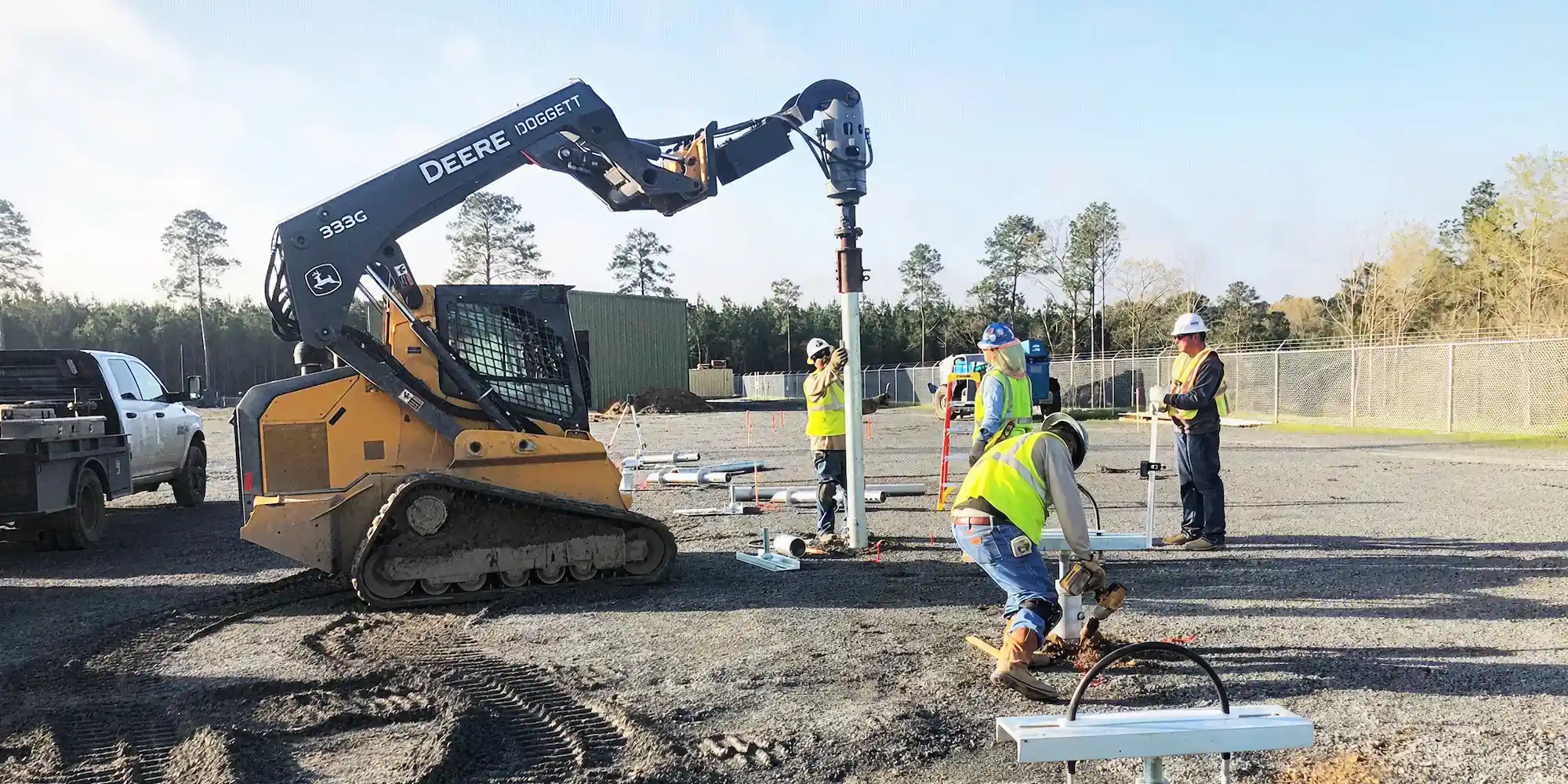 Construction workers in safety vests operating machinery and tools on a gravel site with trees and fencing in the background.