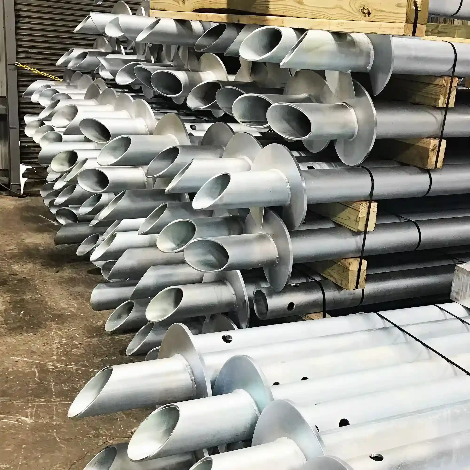 Stack of galvanized steel helical piles with spiral plates for foundation support, held together with wooden blocks and straps.