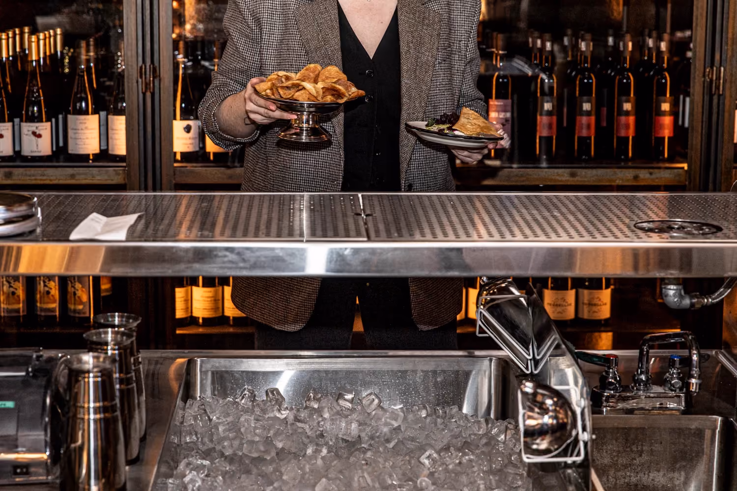 Person behind bar holding a plate of food in one hand and a bowl of chips in the other, with bottles of wine in the background.