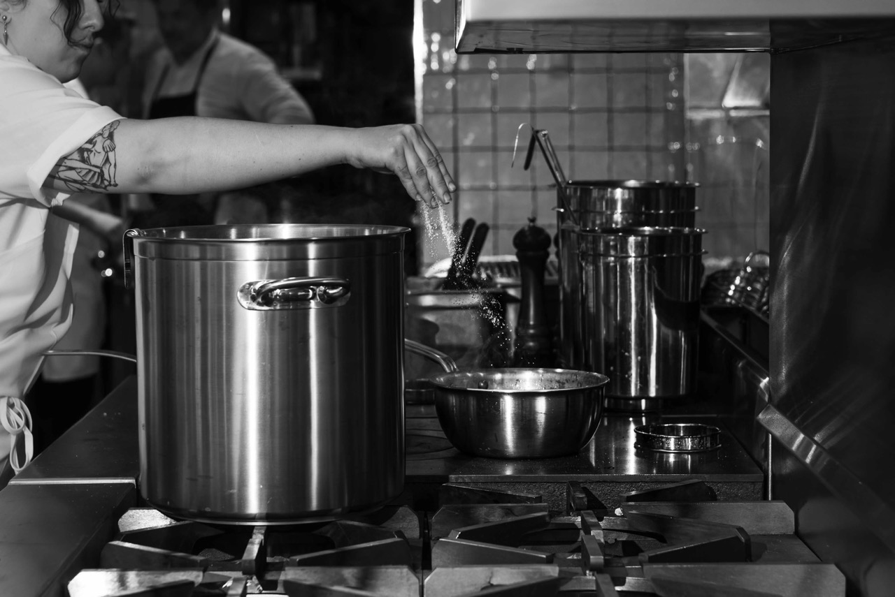 Chef seasoning food by sprinkling salt into a large pot on a professional stove in a kitchen.