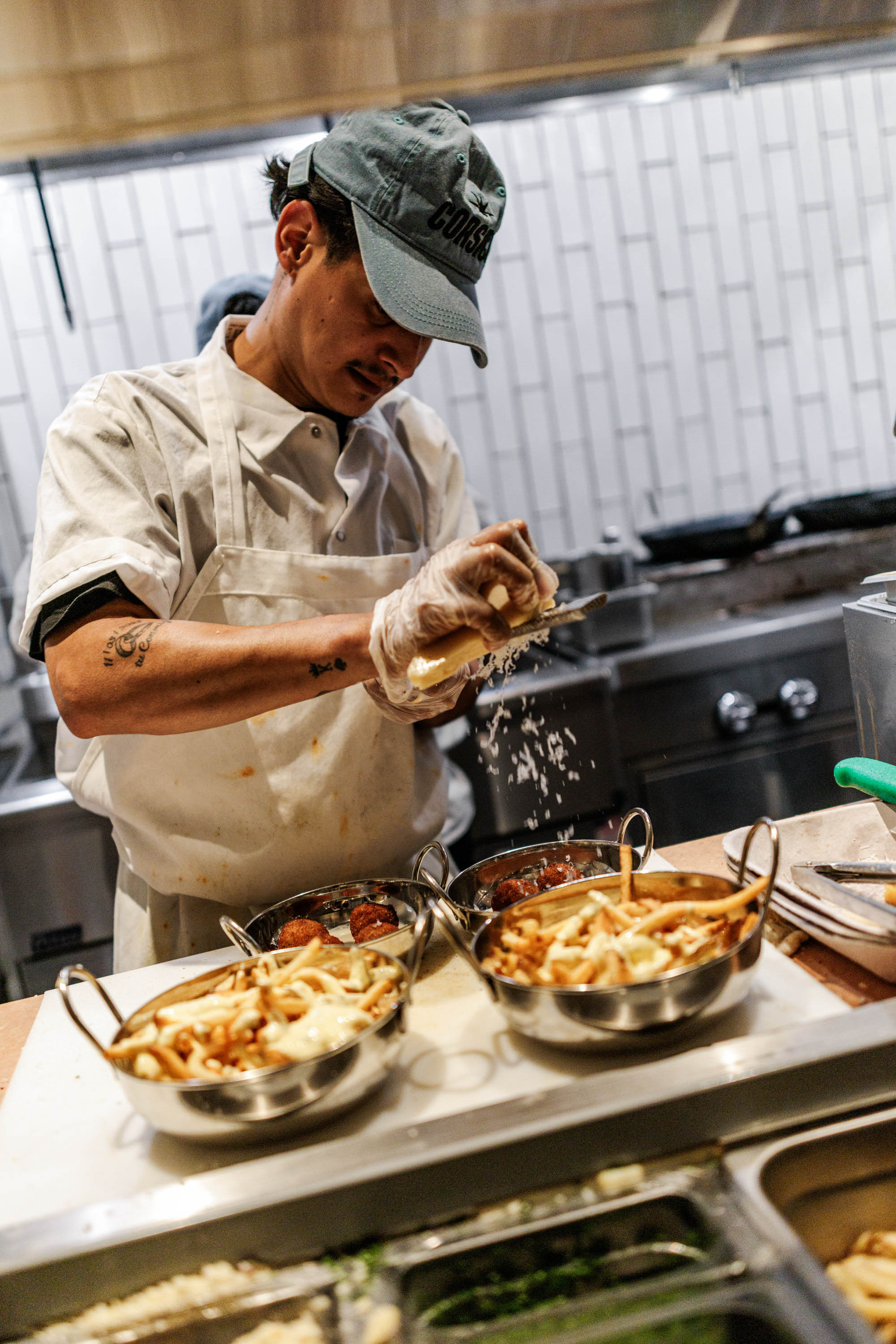 Chef wearing a cap and apron grating cheese over bowls of fries and food in a commercial kitchen.